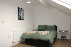 A bedroom with a bed, a radiator, a small table, and a painting on the wall. - Stylish Bradford Home | Near City Centre | Sleeps 11 (Bradford)