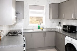 A modern kitchen with grey cabinets, a gas stove, a microwave, and a washing machine. - Contractors or Families | Sleeps 12 + Fast WiFi | Near Bradford University + BRI (Bradford)