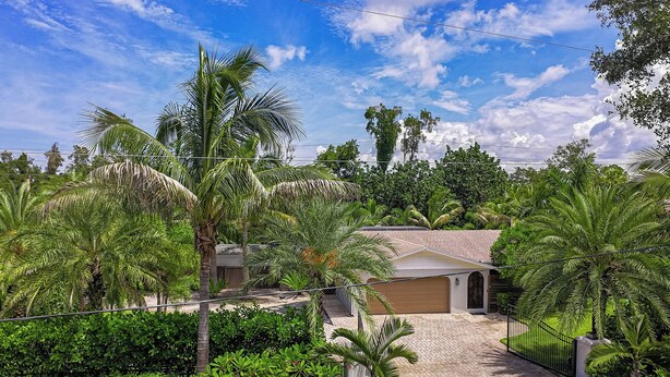 A residential property with a paved driveway leading to a garage, surrounded by lush greenery and palm trees. - Heated Pool & Spa • 3 BRs • 10 min to Siesta Key (Sarasota)