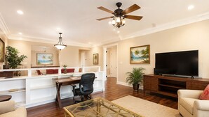 A spacious living room with a glass coffee table, a sofa, a TV, and a ceiling fan. - Beachfront 6BR, 2 Condos for 16 at Turtle Bay! (Kahuku)