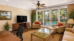A living room with a television, seating, a glass coffee table, and a view of a pool and palm trees. - Beachfront 6BR, 2 Condos for 16 at Turtle Bay! (Kahuku)
