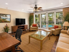 Ein Wohnzimmer mit Fernseher, Sitzgelegenheiten, einem Glastisch und Blick auf einen Pool und Palmen.