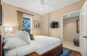 A hotel room with a bed, a ceiling fan, a TV mounted on the wall, a window with curtains, and a door leading to another room.