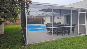 Un espace extérieur aménagé, protégé par des moustiquaires, avec une piscine, du mobilier de jardin et une vue sur une maison au loin.