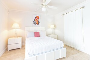 A bedroom with a bed, two bedside tables, a ceiling fan, a wall-mounted artwork, and a window with curtains. - Caprice 5 - 3BR Oceanfront Villa + Pool (Nassau)