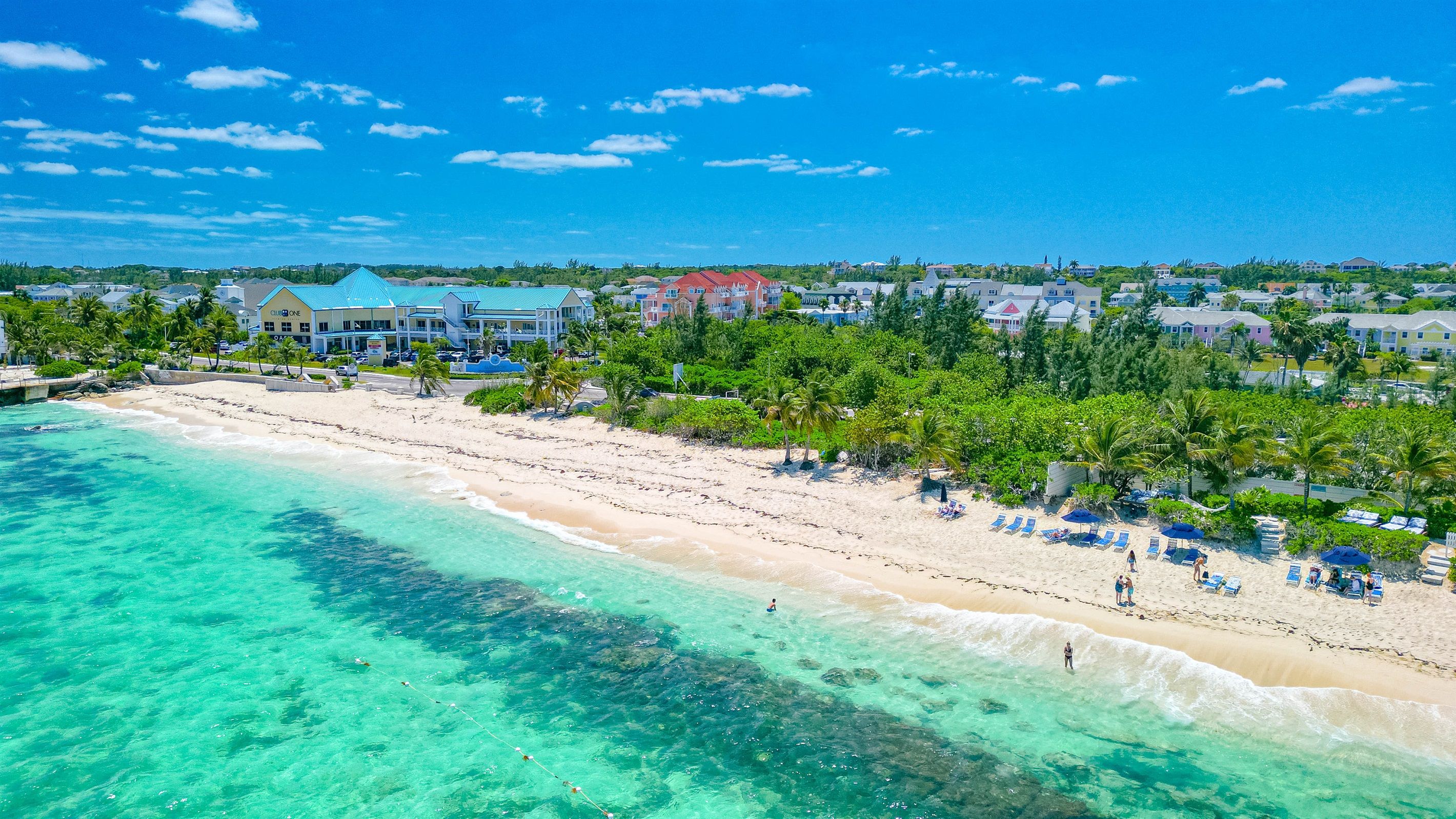 A beach with clear turquoise waters, a sandy shore, and a row of beachside buildings with a row of palm trees in the background.
