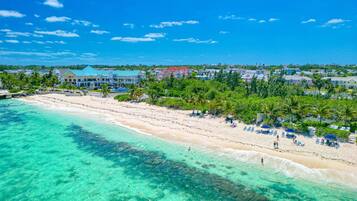A beach with clear turquoise waters, a sandy shore, and a row of beachside buildings with a row of palm trees in the background.