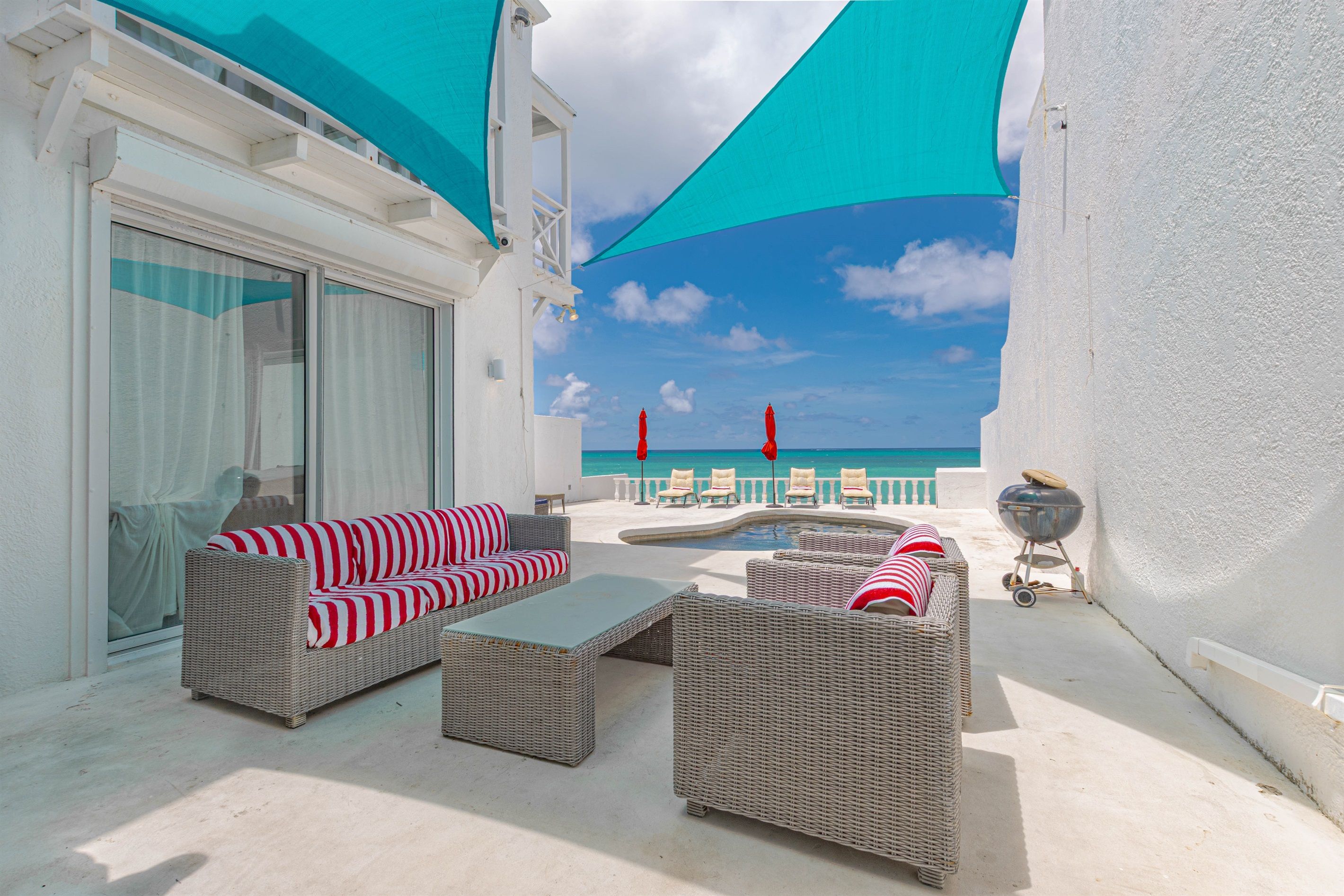 A sunlit outdoor seating area with wicker furniture, a barbecue grill, and a view of the ocean.