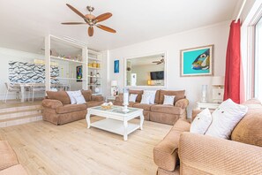 A spacious living room with two sofas, a coffee table, and a ceiling fan. There is a bookshelf and a dining area in the background. - Caprice 5 - 3BR Oceanfront Villa + Pool (Nassau)