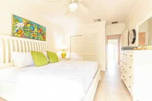 A bedroom with a bed, a nightstand, a lamp, a dresser, a mirror, a painting, and a door leading to another room. - Caprice 5 - 3BR Oceanfront Villa + Pool (Nassau)