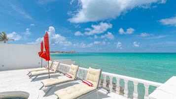 A poolside area with lounge chairs and a red umbrella, overlooking a clear blue sea.