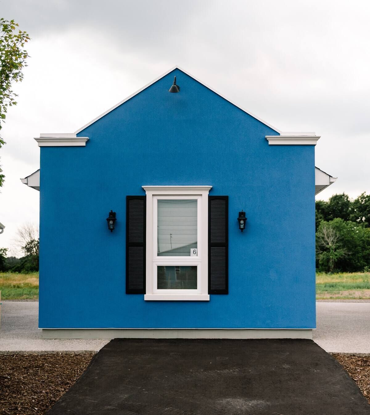 Ein kleines, eingeschossiges Gebäude mit blauer Fassade, einem Fenster mit weißem Rahmen und Fensterläden sowie zwei Wandleuchten.