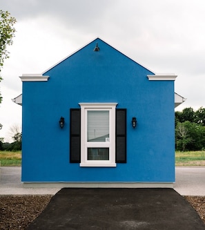 A small, single-story building with a blue exterior, a white-framed window with shutters, and two wall-mounted light fixtures. - New Vineyard Cottage in Niagara Wine Country | 1BR (Lincoln)