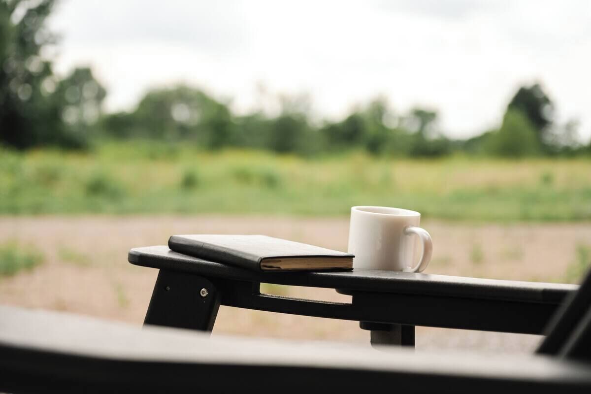 A white mug and a book on a black table with a view of a green field.