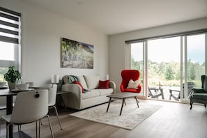 A modern living room with a sofa, chairs, a coffee table, and a large window offering a view of the outdoors. - New Vineyard Cottage in Niagara Wine Country | 1BR (Lincoln)