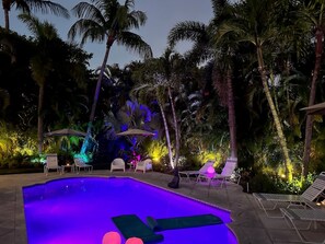 A swimming pool with lounge chairs and umbrellas, surrounded by palm trees and illuminated by ambient lighting. - The Key West House | Tropical Oasis |Close 2 Beach (West Palm Beach)