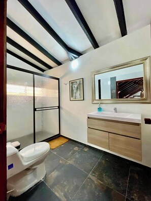 A modern bathroom with a toilet, a shower with frosted glass doors, a vanity with a mirror, and a tiled floor. - Hacienda Familiar - Estancia 30P - Wifi (Ulloa)