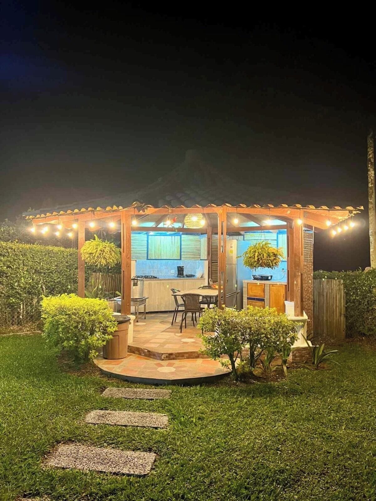 A well-lit outdoor kitchen area with a dining table and chairs, surrounded by a wooden pergola with string lights.