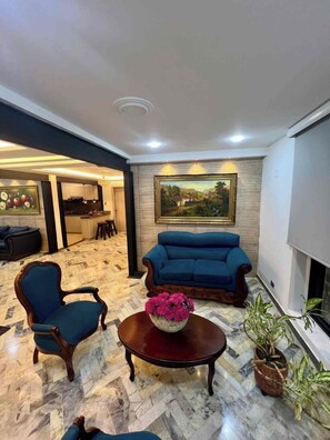 A spacious living area with a sofa, armchair, and coffee table, a painting on the wall, and a kitchen in the background. - Hacienda Familiar - Estancia 30P - Wifi (Ulloa)