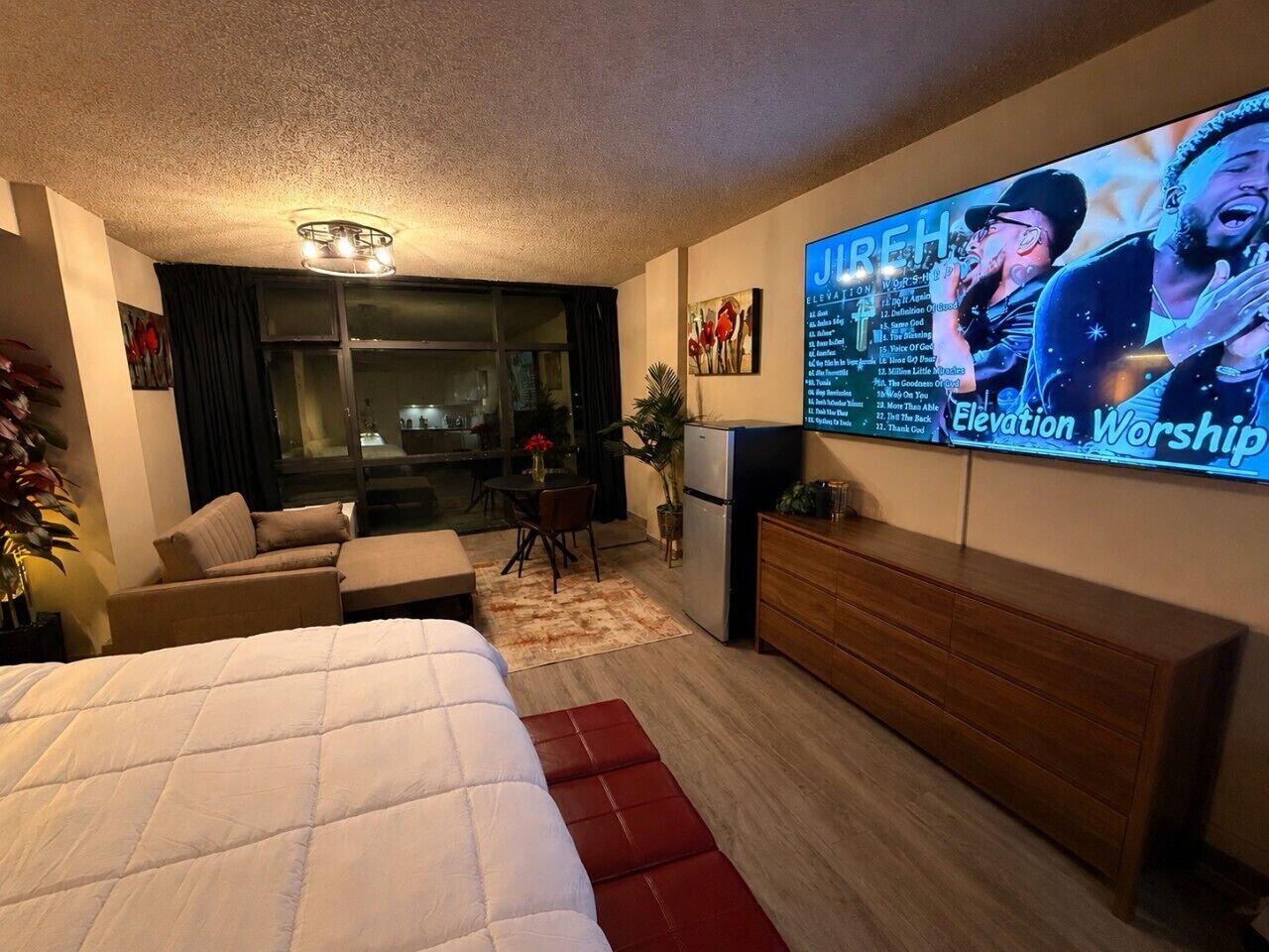 Quarto de hotel com cama, televisão exibindo um culto, sofá, mesa pequena e área de cozinha compacta.