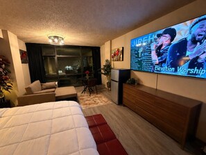Quarto de hotel com cama, televisão exibindo um culto, sofá, mesa pequena e área de cozinha compacta.