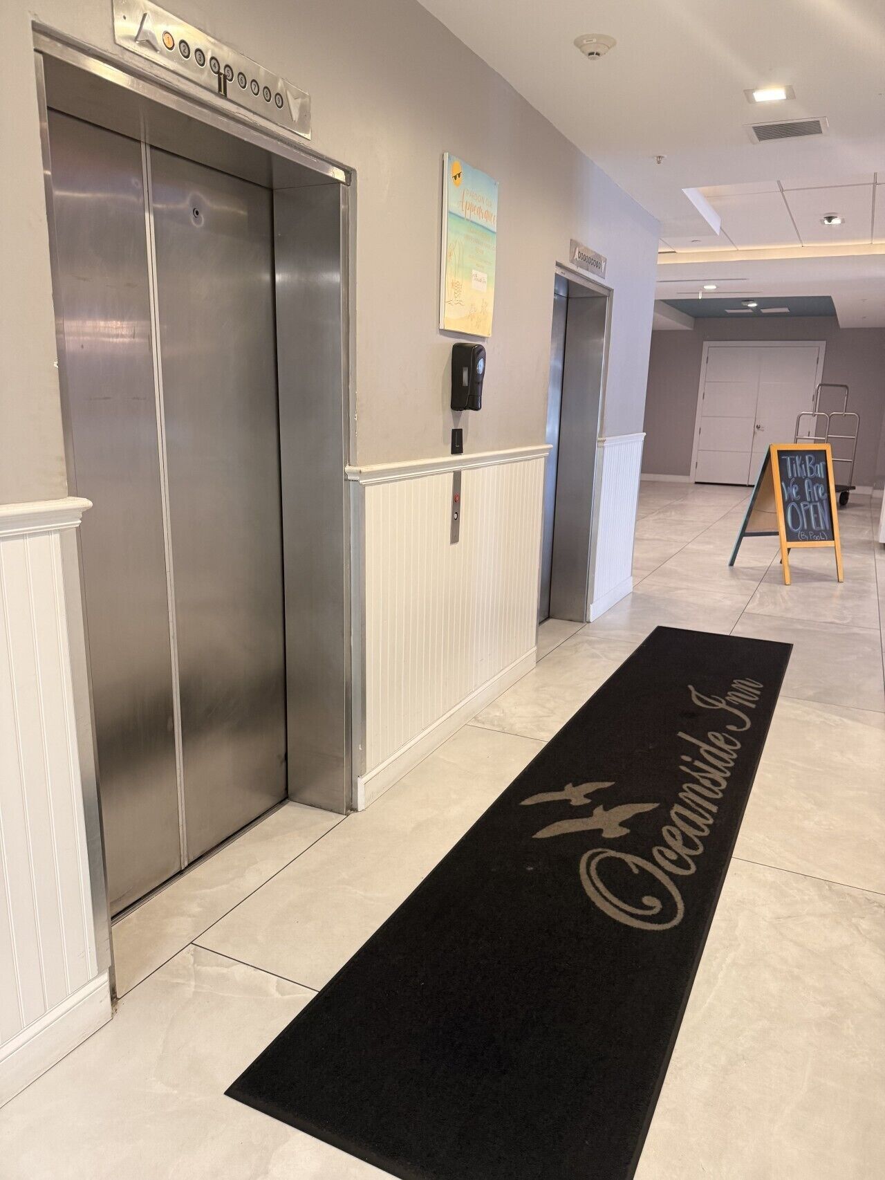 Un hall d'hôtel avec deux ascenseurs, un tapis noir orné du texte « Oceanside Inn », et un panneau indiquant que le bar est ouvert.