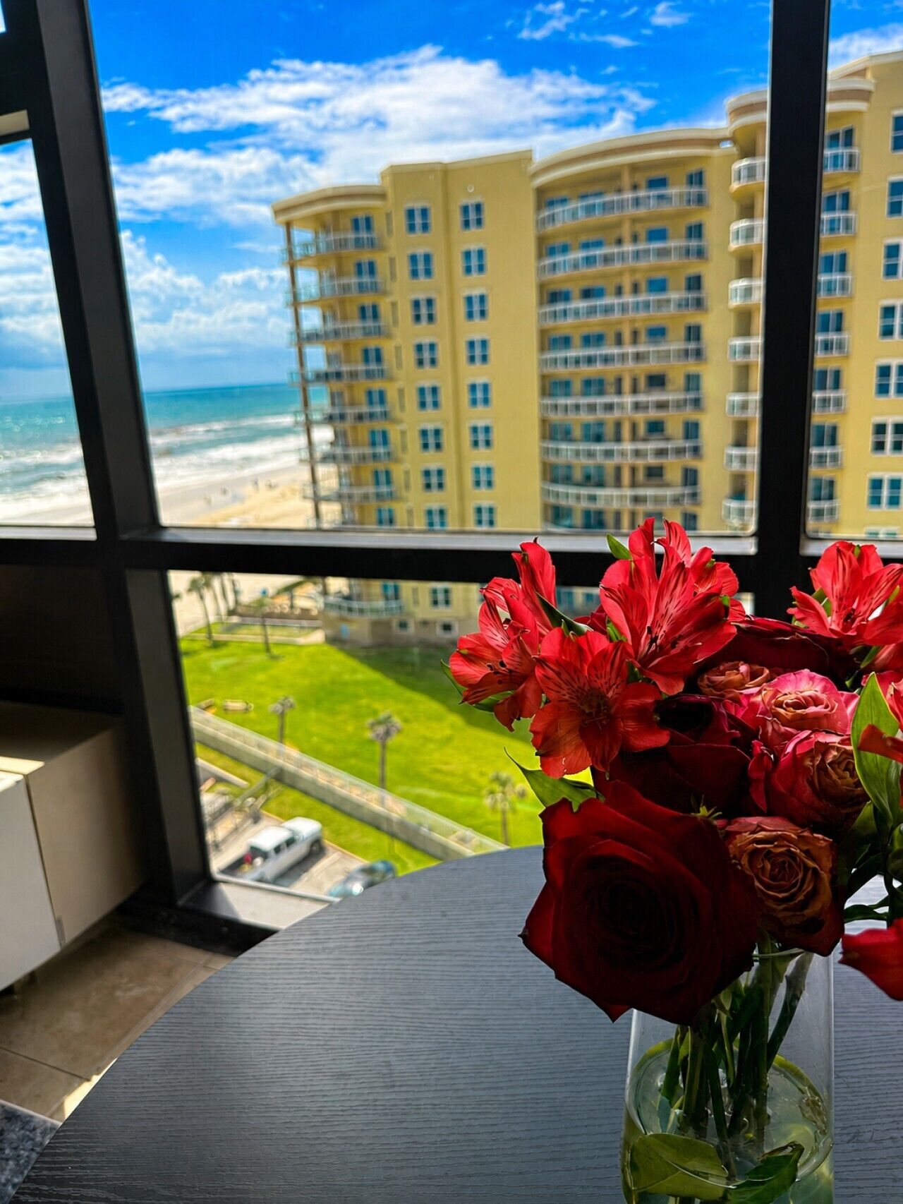 Vista da janela de um edifício amarelo, com uma praia e um vaso de flores vermelhas sobre uma mesa.