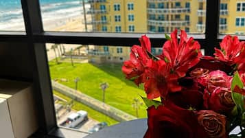 Vista da janela de um edifício amarelo, com uma praia e um vaso de flores vermelhas sobre uma mesa.