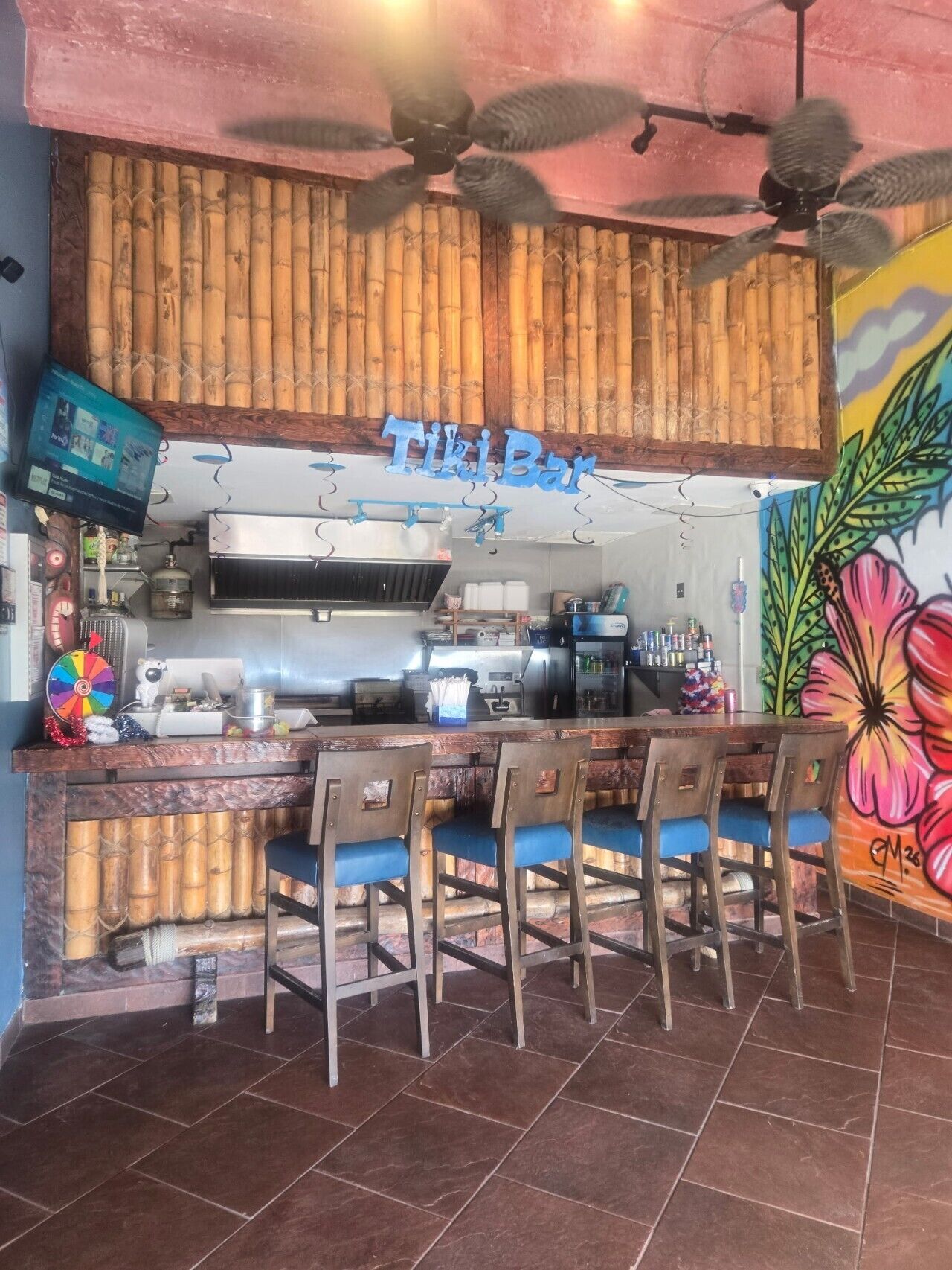 Uma área de bar com teto de madeira, uma placa com a inscrição 'Tiki Bar' e um mural colorido na parede.