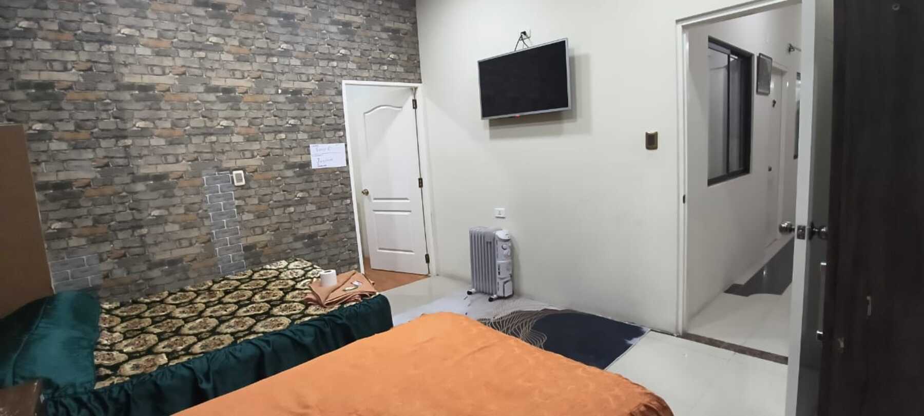 A bedroom with a brick wall, a bed with patterned bedding, a radiator, a television, and a door.