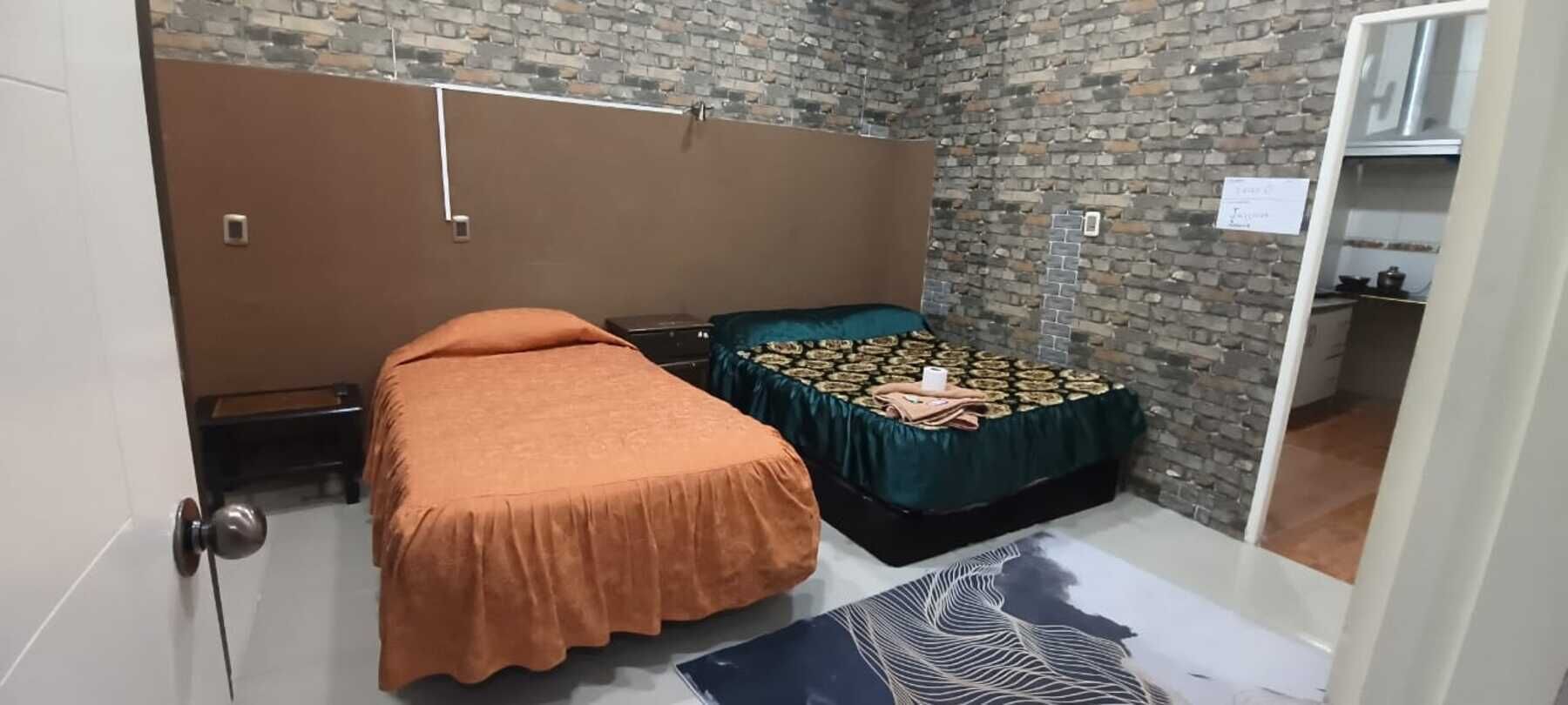 A small, compact room with a brick wall, a bed with an orange bedspread, a bench, and a small table with a green tablecloth.
