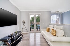 Ein modernes Wohnzimmer mit einem Flachbildfernseher, einem Stativ, einem weißen Sofa mit goldenen Kissen und einer Schiebetür zu einem Außenbereich.