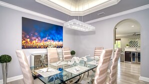A modern dining room with a glass table, cushioned chairs, a large abstract painting, and a crystal chandelier. - Private Oasis - Pool & Backyard (Hialeah)