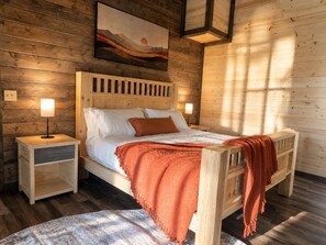 A wooden cabin bedroom with a bed, nightstands, a painting, and a window allowing sunlight. - Luxury Smoky Retreat | Pool, Theater & Hot Tub (Sevierville)