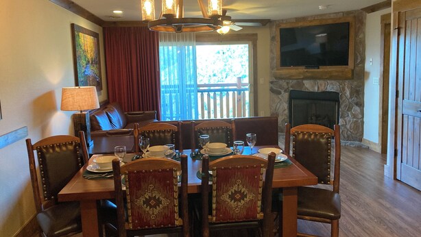 A dining area with a wooden table set for four, surrounded by upholstered chairs with decorative backs. A chandelier hangs from the ceiling, and a stone fireplace is visible in the background. - Cozy 1-bedroom condo with balcony in beautiful Gatlinburg with Water Park passes (Gatlinburg)