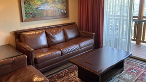 A living room with a brown leather sofa, a wooden coffee table, and a large painting of a river scene. - Cozy 1-bedroom condo with balcony in beautiful Gatlinburg with Water Park passes (Gatlinburg)