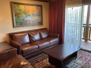 A living room with a brown leather sofa, a wooden coffee table, and a large painting of a river scene. - Cozy 1-bedroom condo with balcony in beautiful Gatlinburg with Water Park passes (Gatlinburg)