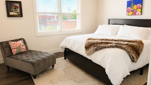 A bedroom with a bed, a chair, a window, and two paintings.