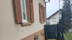 A building with a beige exterior, wooden shutters on windows, a metal gate, and a paved area with moss. - Vacation Home in a Rural Setting (Heidwiller)