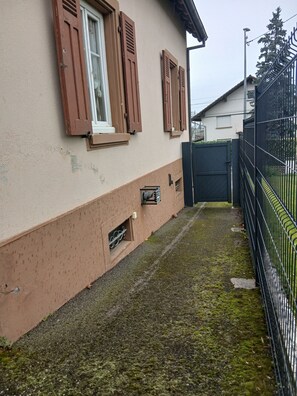 A building with a beige exterior, wooden shutters on windows, a metal gate, and a paved area with moss.