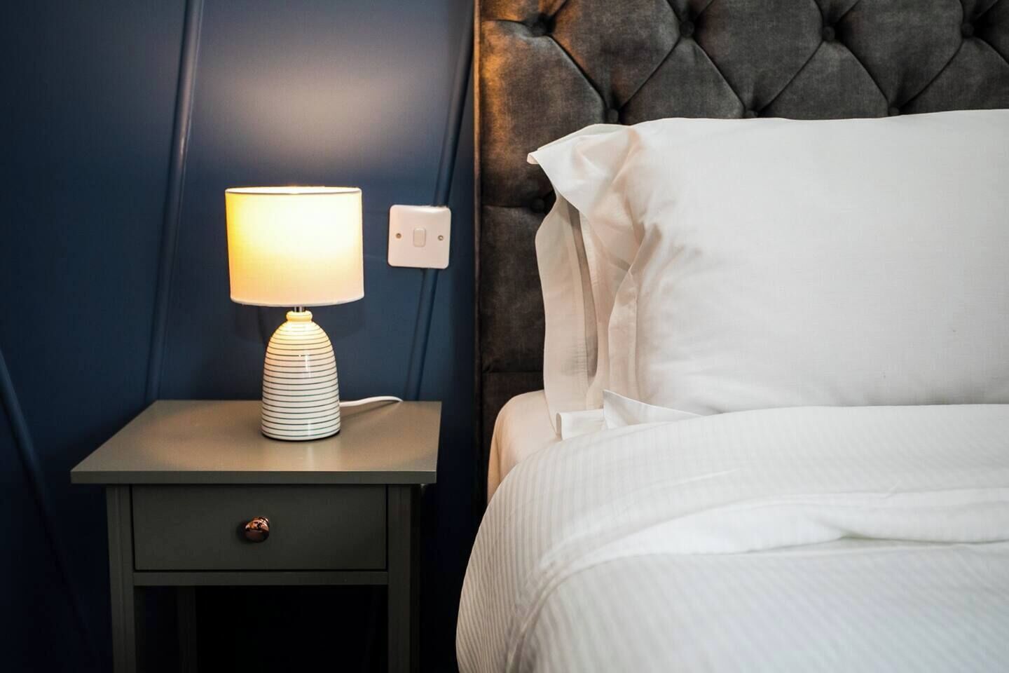 A neatly made bed with white linens, a bedside lamp with a striped base, and a dark blue wall.
