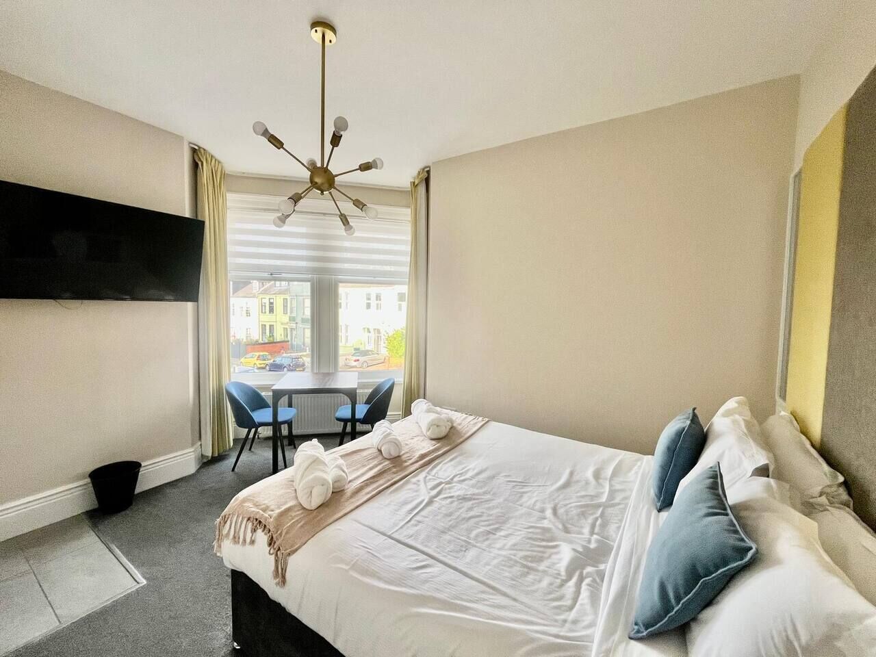 A hotel room with a bed, a TV mounted on the wall, a dining table with chairs, and a view of the street through the window.