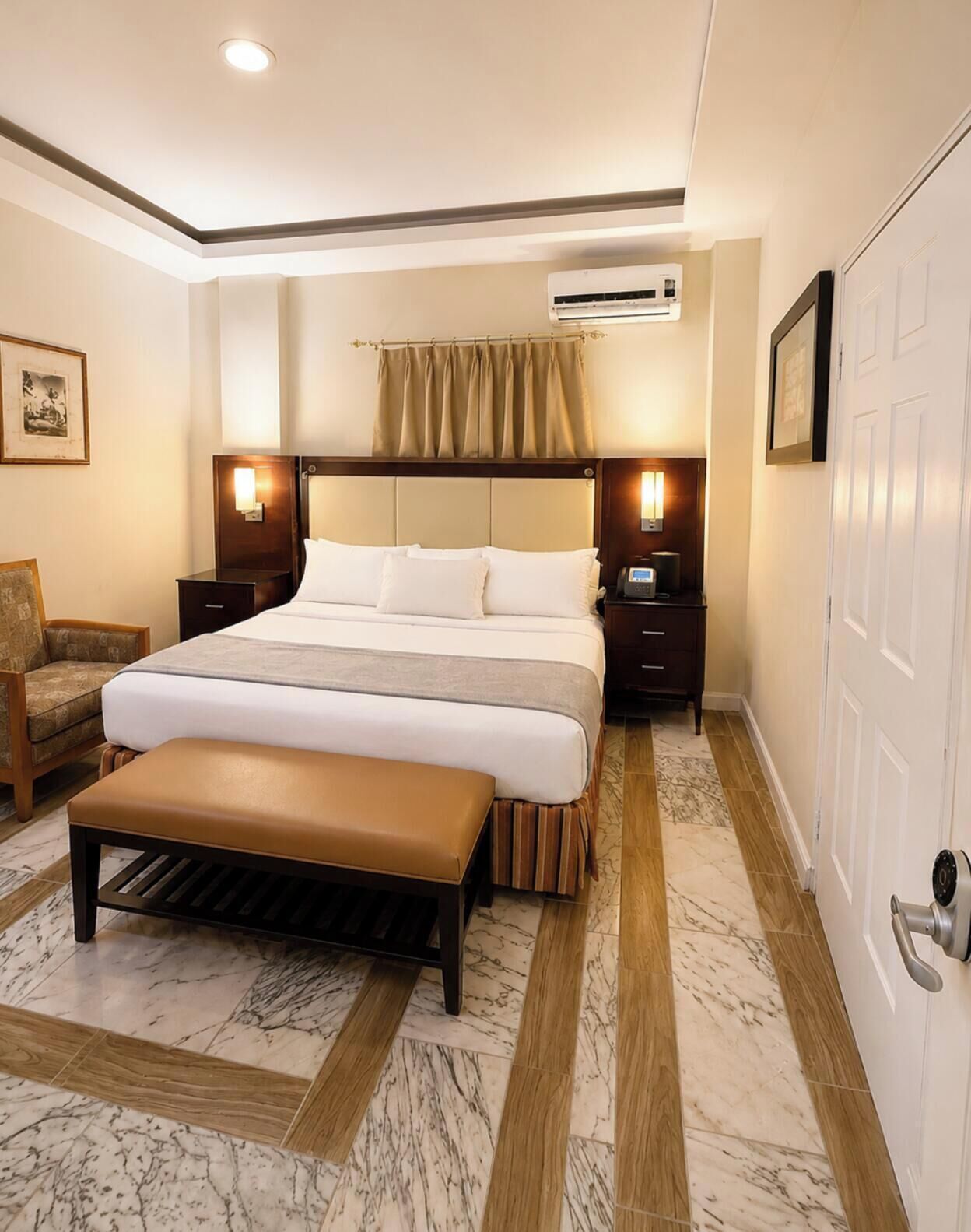 Un dormitorio de hotel con cama, mesitas de noche, equipo de aire acondicionado, un banco y una puerta.