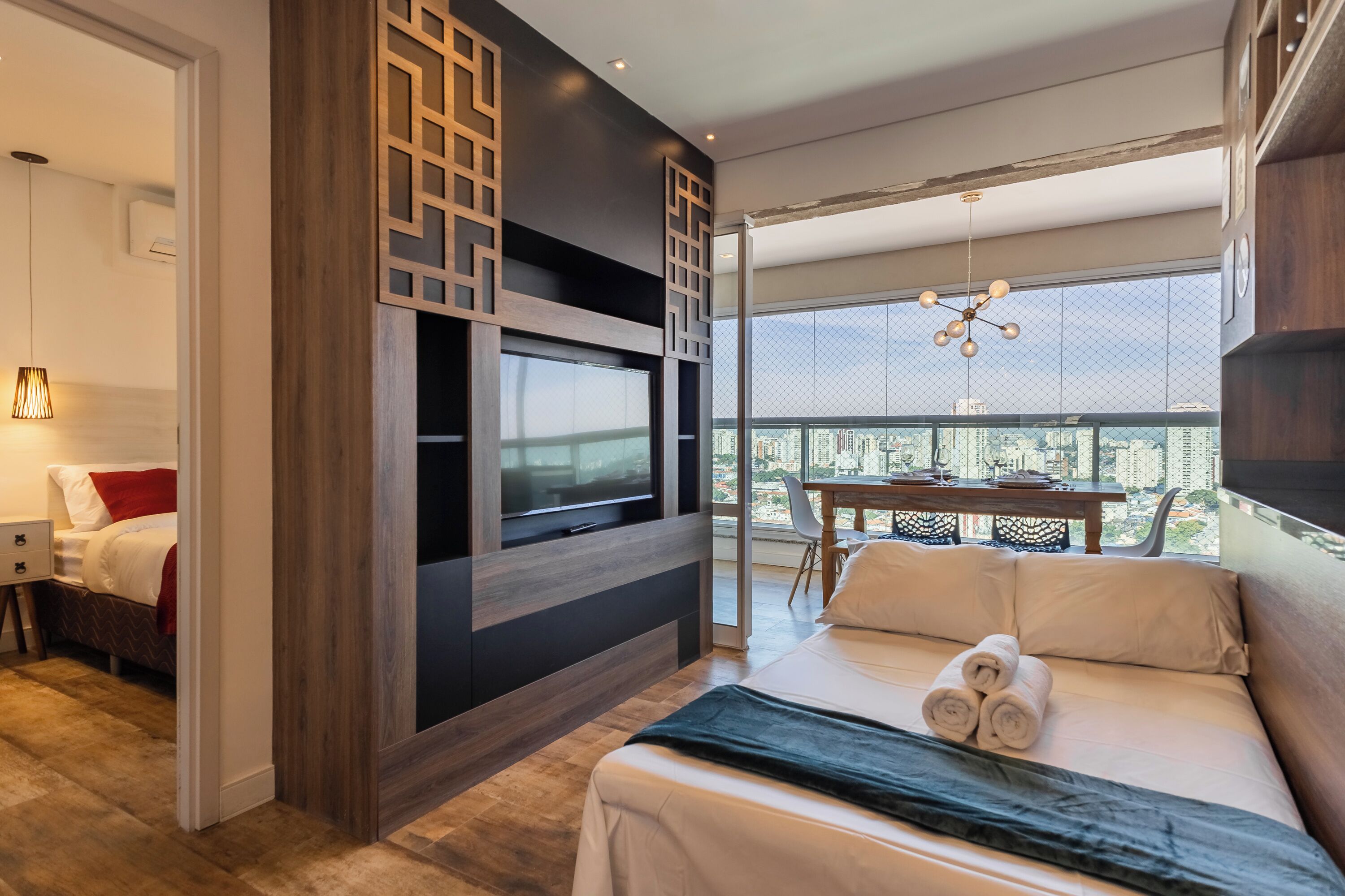 Ein modernes Hotelzimmer mit einem großen Bett, einem an der Wand befestigten Fernseher, einem Balkon mit Tisch und Stühlen sowie Blick auf die Stadt.