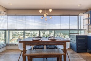 A dining area with a table set for two, a modern chandelier, and a view through a large window with a metal fence. - StayHome - Campo Belo - Gym & Pool (São Paulo)