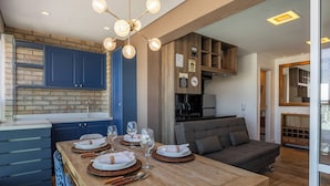 Una cocina moderna con armarios azules, una zona de comedor con una mesa de madera preparada para cuatro personas, y un sofá gris en la sala de estar.