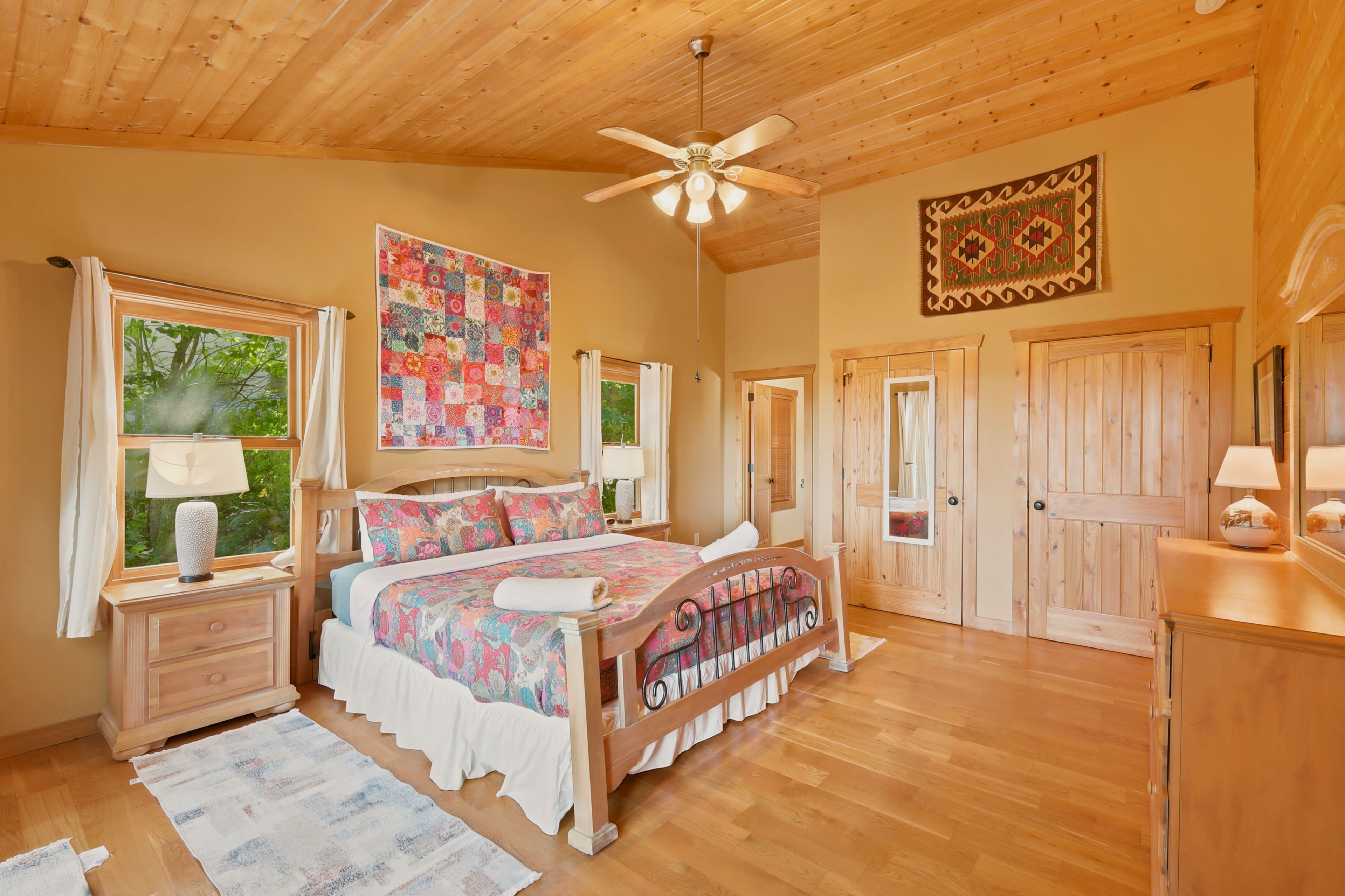 Spacious bedroom with a king bed in a wood and wrought iron frame, floral quilt and rolled towels, nightstands with lamps, a patchwork quilt and woven rug on the walls, two windows to trees, two closet doors, dresser and ceiling fan.