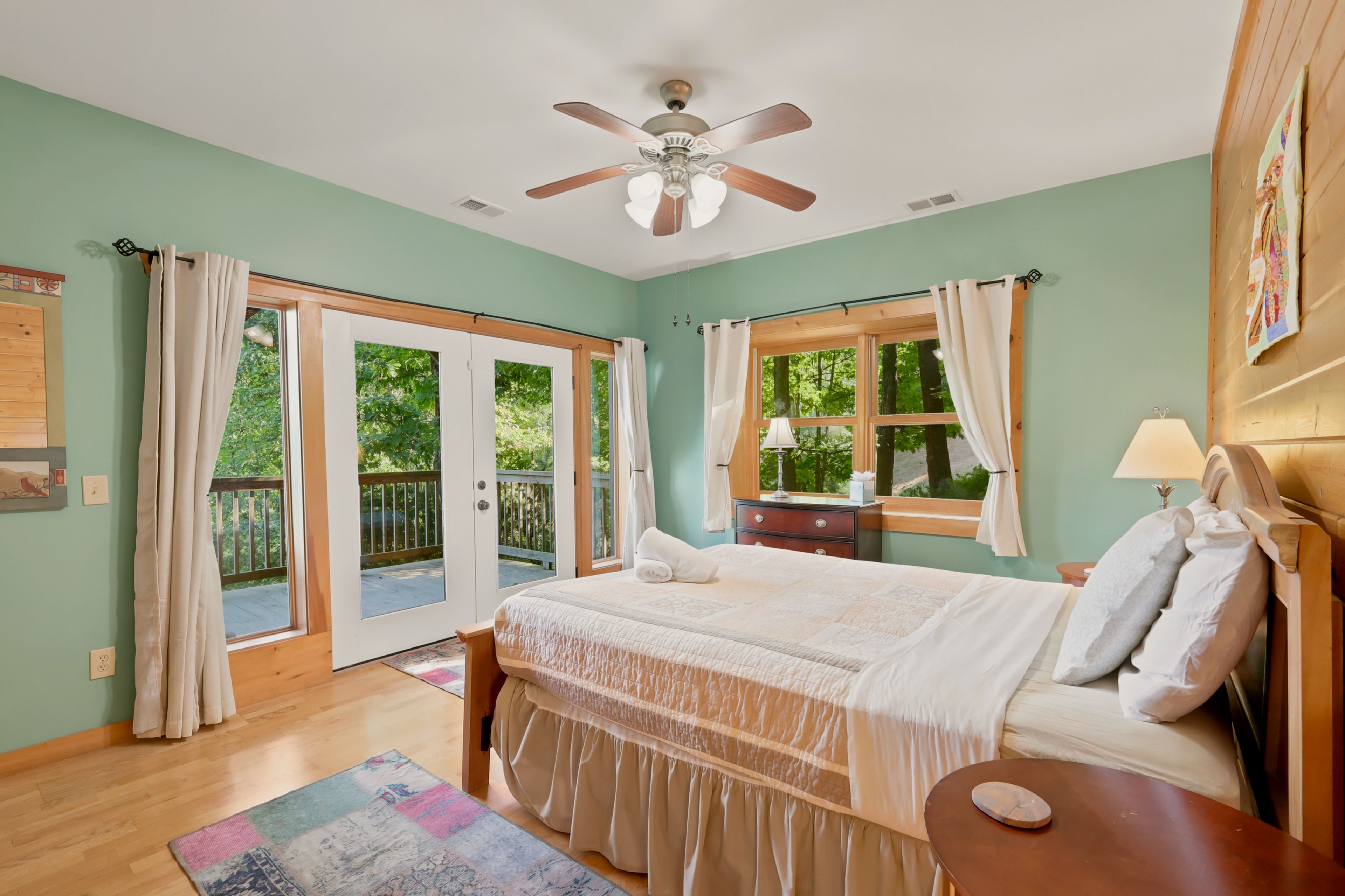A bedroom with a bed, a nightstand, a ceiling fan, a window with curtains, and a view of the outdoors.