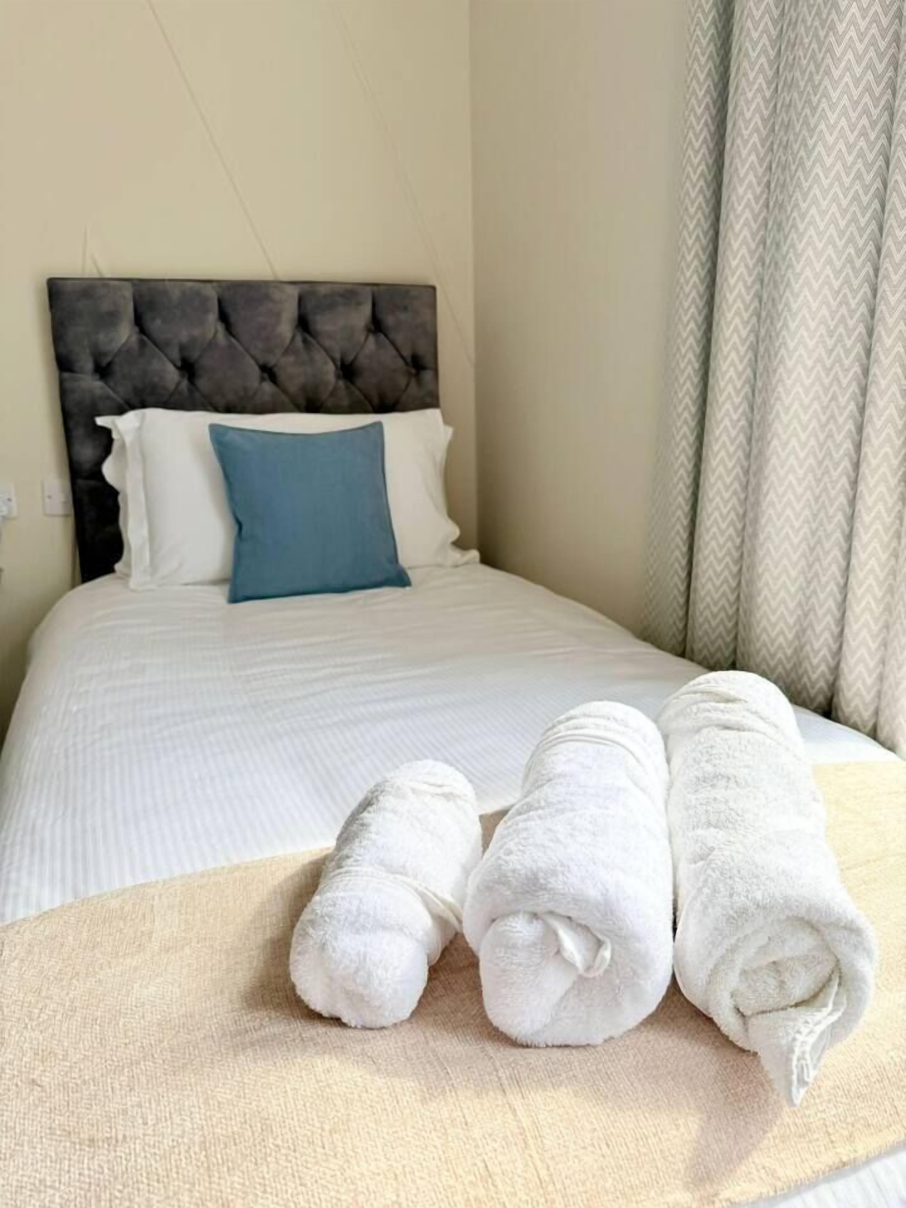 A neatly made bed with a tufted headboard, a blue pillow, and rolled white towels on a beige bedspread.