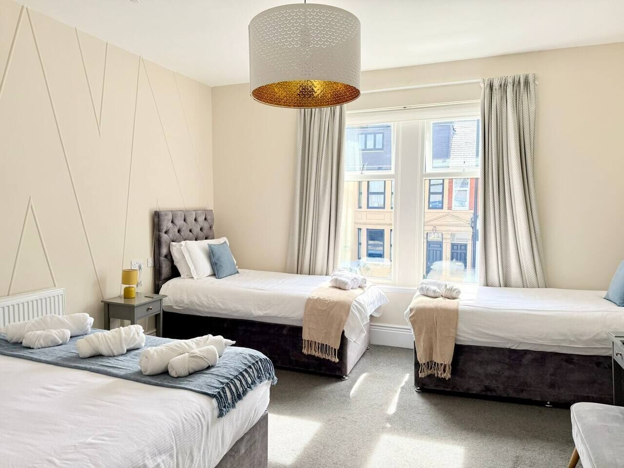 Ett hotellrum med två sängar, ett stort fönster med gardiner, en hängande lampa och ett element.
